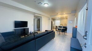 A modern living room with a dark-colored sofa, a flat-screen TV, and a small coffee table. There is a dining area with a round table and chairs, and a kitchen visible in the background.