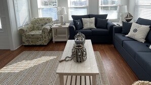 A living room with a ceiling fan, a sofa set, a patterned armchair, a coffee table, and a view of the water through large windows. - Redecorated 3 Bed ,2 Bath ,Sofa bed Outdoor shower Come relax at our beach house (Surfside Beach)