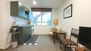 A compact kitchen with a microwave, kettle, and washing machine. There is a dining area with a table and chairs, and a window with a view of buildings outside. - City Living - Wifi- Netflix - Laundry (Auckland)