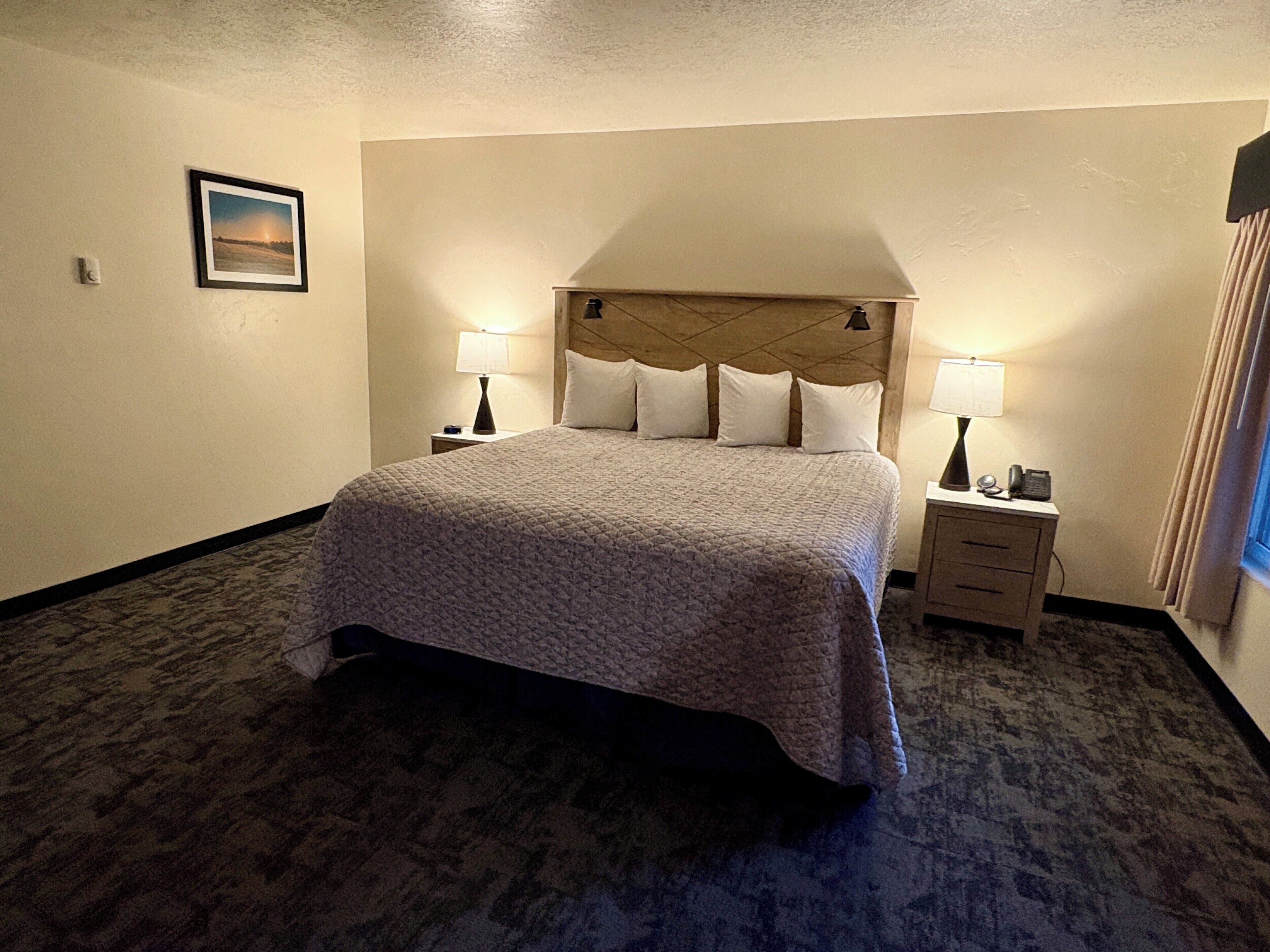 A hotel room with a large bed, two bedside tables with lamps, a headboard, and a framed picture on the wall.