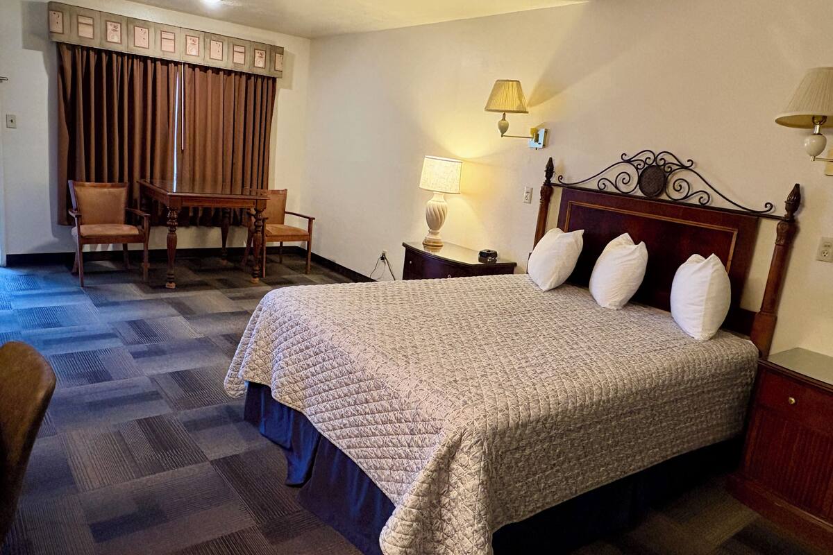 A hotel room with a large bed, a desk, two chairs, a lamp, and a wall clock.