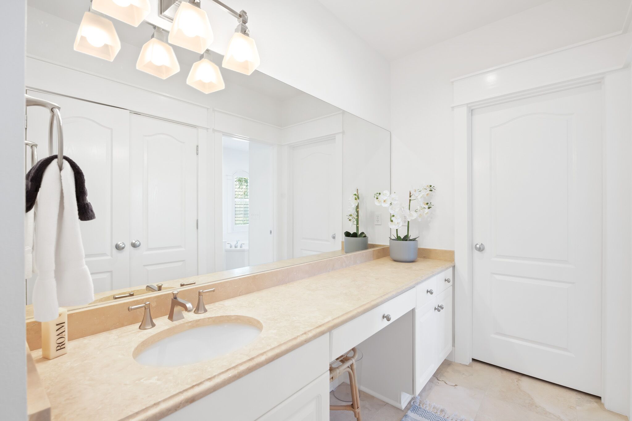 A modern bathroom with a beige countertop, a large mirror, and a sink. There are white cabinets and a door visible.