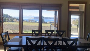 A dining area with a large table and chairs, large windows with a view of a lake and mountains, and a door leading outside.
