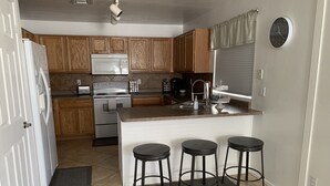 A kitchen with wooden cabinets, a white refrigerator, a microwave, a stove, a sink, and a clock on the wall. There are three stools at the island. - Fun 4 bedroom house w/ splash pad, near City Center and Saguaro National Forest (Tucson)