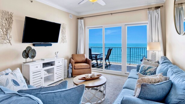 A spacious living room with a flat-screen TV, a ceiling fan, a sofa set, a coffee table, and a view of the ocean through large sliding doors. - Seawind 1005 | Beautiful condo with perfect beach view and walk to dine, shop, more! (Gulf Shores)