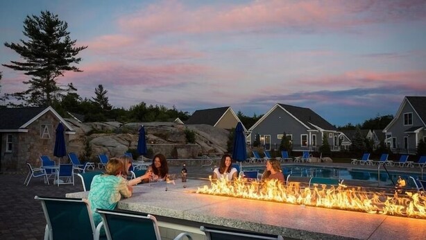 A poolside area with a fire feature, outdoor seating, and a view of houses in the background. - Sunkissed cottage, heated pools, club house, game room, 15 mins to local beaches (Arundel)