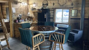 A rustic cabin interior with a round wooden table surrounded by chairs, a kitchen area with a refrigerator, and a window with a view of the outdoors.