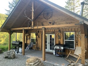 Een rustieke blokhut met een overdekte veranda, een barbecue en een grote rots op de voorgrond.