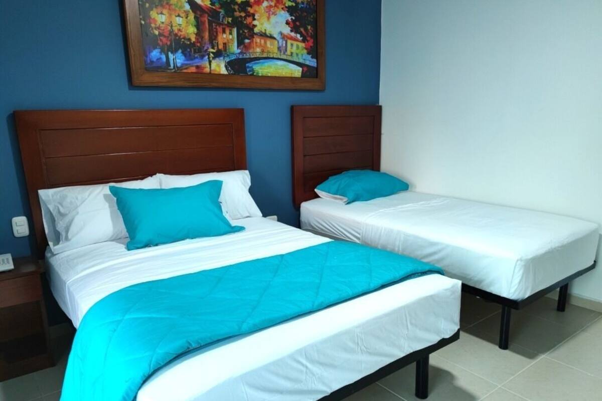 A hotel room with two beds, each with white bedding and a teal accent pillow. A painting of a cityscape hangs above the beds.