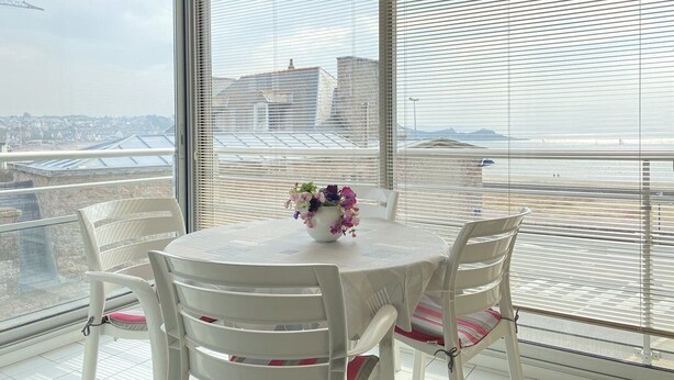 Ein Balkon mit rundem Tisch und Stühlen, einem Blumenstrauß und Blick auf Gebäude sowie eine Brücke.