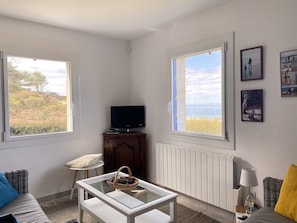 Una habitación con sofá, una mesita, televisión y ventana con vista al mar.