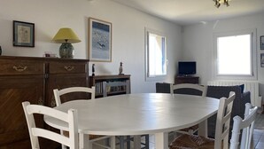Ruokailutila, jossa on valkoinen pöytä ja tuolit, puinen kaappi, lamppu ja seinillä olevia kehystettyjä taideteoksia.