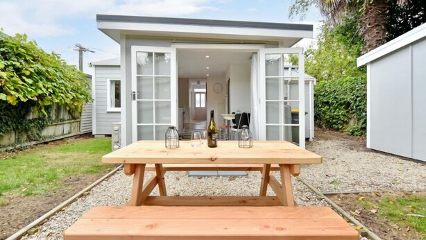 Una mesa de picnic de madera con una botella y vasos en un patio trasero con un cobertizo y una casa al fondo.