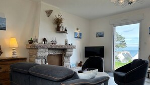 A living room with a stone fireplace, a television, and a view of the sea. - Sea-view house in Erquy | 8 guests | Ref 70 (ERQUY)