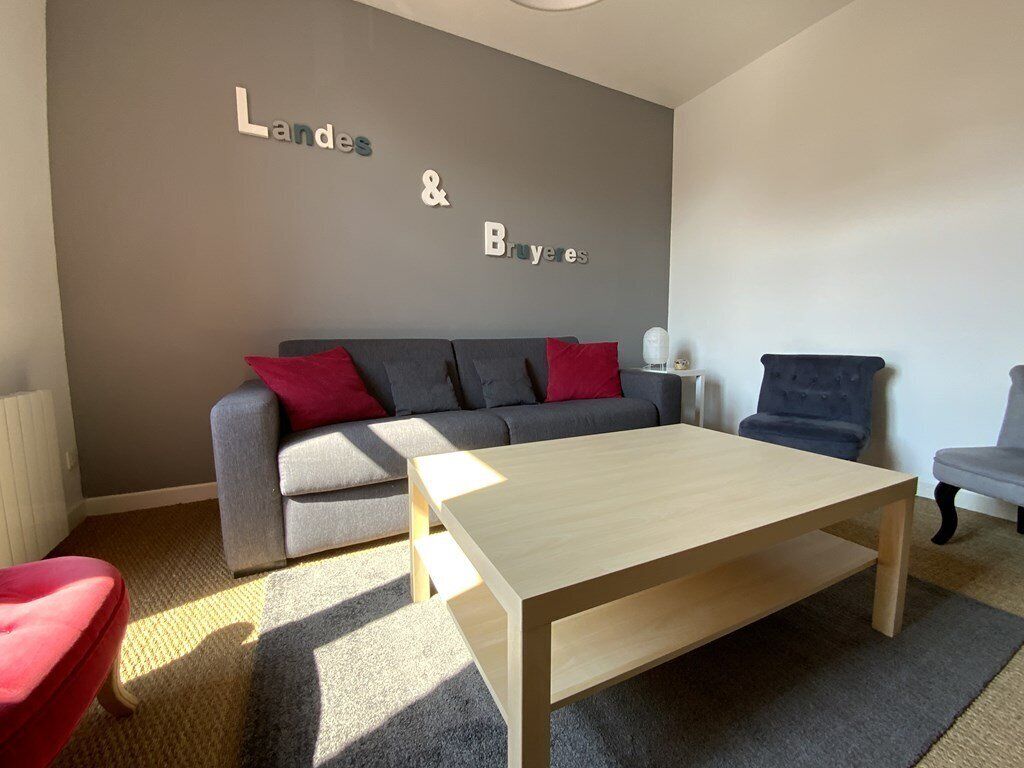 A modern living room with a grey sofa, red and black cushions, a wooden coffee table, and a wall-mounted sign reading "Landes & Bruyères".