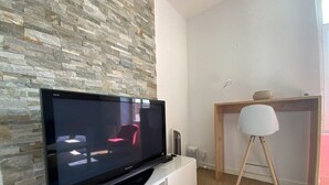 A modern living room with a flat-screen TV on a white entertainment unit, a stone wall, a wooden desk with a white chair, and a carpeted floor. - Apartment near beach in Erquy | 5 guests | Ref 653 (ERQUY)