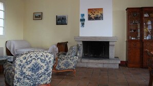 Een woonkamer met een open haard, twee gestoffeerde fauteuils, een houten kast en kunstwerken aan de muur.