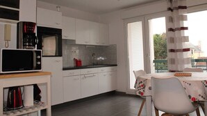 Une cuisine compacte avec des meubles blancs, un four à micro-ondes et une machine à café. On y trouve un coin repas avec une table et des chaises, ainsi qu’une fenêtre avec des rideaux.