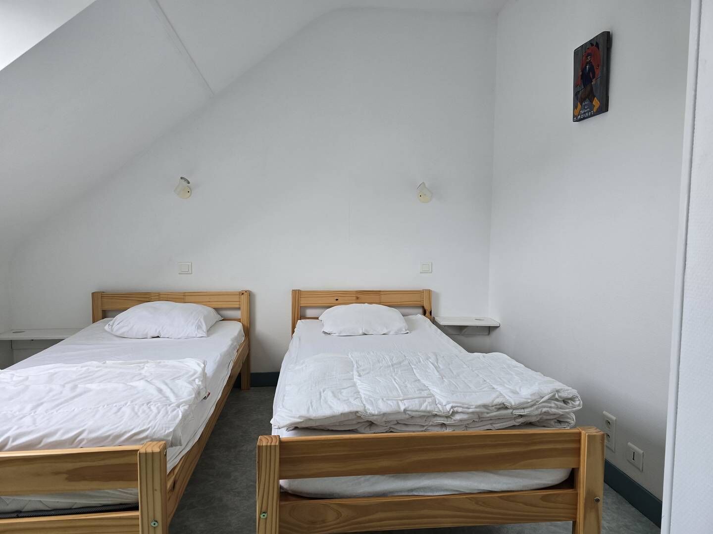 A dormitory room with two wooden bunk beds, white bedding, and a small shelf on the wall.