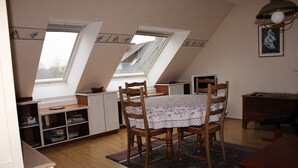 A room with a dining table, chairs, a bookshelf, and a desk. There are two windows providing natural light.