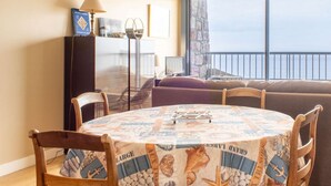 A dining area with a round table covered in a newspaper print tablecloth, surrounded by wooden chairs. A sofa and a small TV are visible in the background, with a stone wall and a view of the sea through a large window. - Sea-view apartment in Erquy 50 m from the beach | 4 guests | Ref 411 (ERQUY)