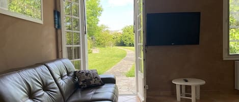 A living room with a leather sofa, a small white table, a TV mounted on the wall, and a door leading to a garden.