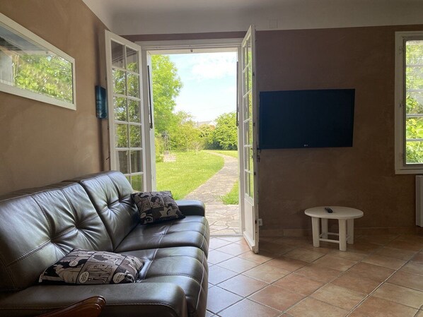 Een woonkamer met een leren bank, een klein wit tafeltje, een aan de muur bevestigde televisie en een deur die uitkomt op een tuin.
