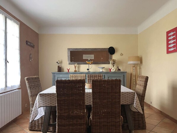 A dining area with wicker chairs, a table with a patterned tablecloth, a blue cabinet with decorative items, and a wall-mounted picture.