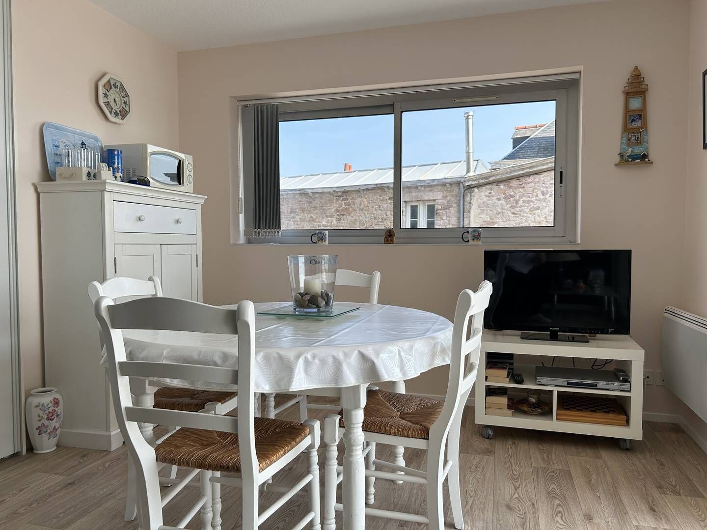 Una habitación con una mesa redonda, sillas blancas, un televisor sobre un mueble bajo, una ventana con vistas a edificios y un armario con utensilios de cocina.
