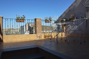 Una terrassa al terrat amb taula i cadires metàl·liques, plantes en testos i vista cap a edificis al fons.