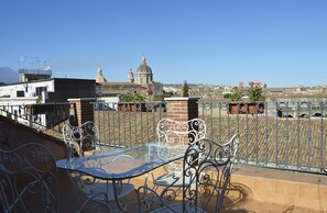 Una terraza en la azotea con una mesa de vidrio y sillas metálicas, con vistas a un paisaje urbano con edificios históricos y un cielo despejado.