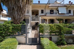 A two-story building with a gated entrance, a balcony on the upper floor, and a small garden area with shrubs.