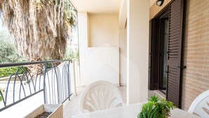 A balcony with a white table and chairs, a potted plant, and a view of a tree and building.