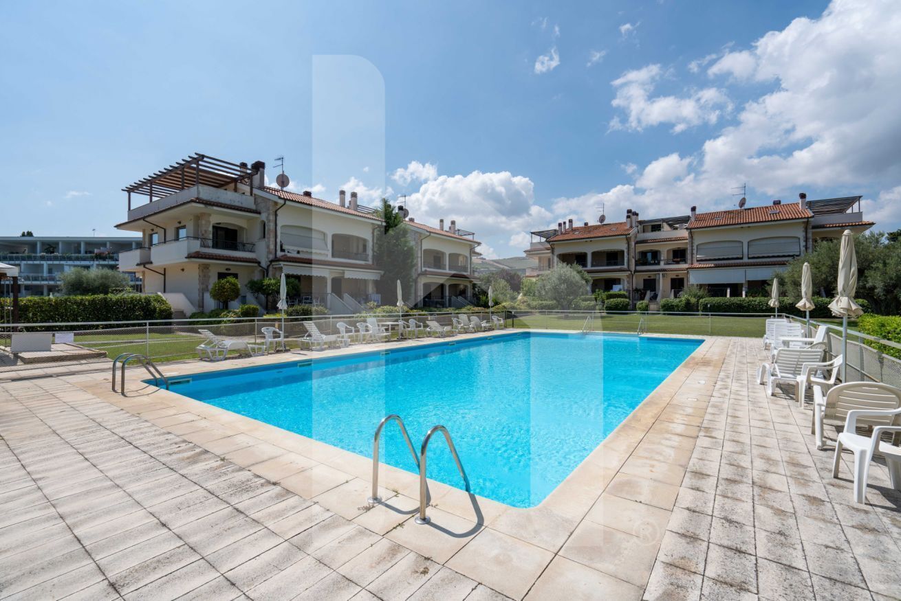 Une piscine dotée d’une terrasse carrelée, entourée de chaises longues et bordée d’un bâtiment comportant plusieurs balcons.