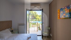 A bedroom with a bed, a nightstand, a lamp, a radiator, a window, and a door leading to an outdoor area with a pool and greenery.