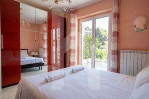 A bedroom with a bed, a wardrobe, a mirror, a window with curtains, and a view of greenery outside.