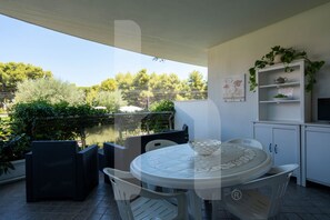 A covered outdoor patio with a round table and chairs, a white cabinet with shelves, and a view of trees and a body of water.