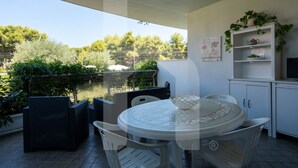 Une terrasse extérieure couverte, avec une table ronde et des chaises, un meuble blanc avec des étagères, et une vue sur des arbres et une étendue d’eau.