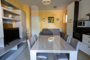 A modern living room with a flat-screen TV, a dining table with chairs, a sofa, and a kitchen area with appliances.