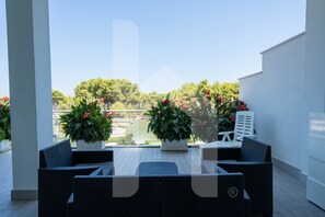 An outdoor patio with wicker furniture, potted plants, and a view of a body of water and trees.