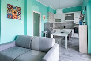 Un salon moderne avec un canapé gris, une table à manger avec des chaises, une cuisine avec des meubles blancs et une peinture abstraite colorée accrochée au mur.