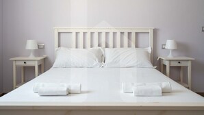Een net opgemaakt bed met witte lakens en kussens, twee nachtkastjes met lampen, en een effen muur.