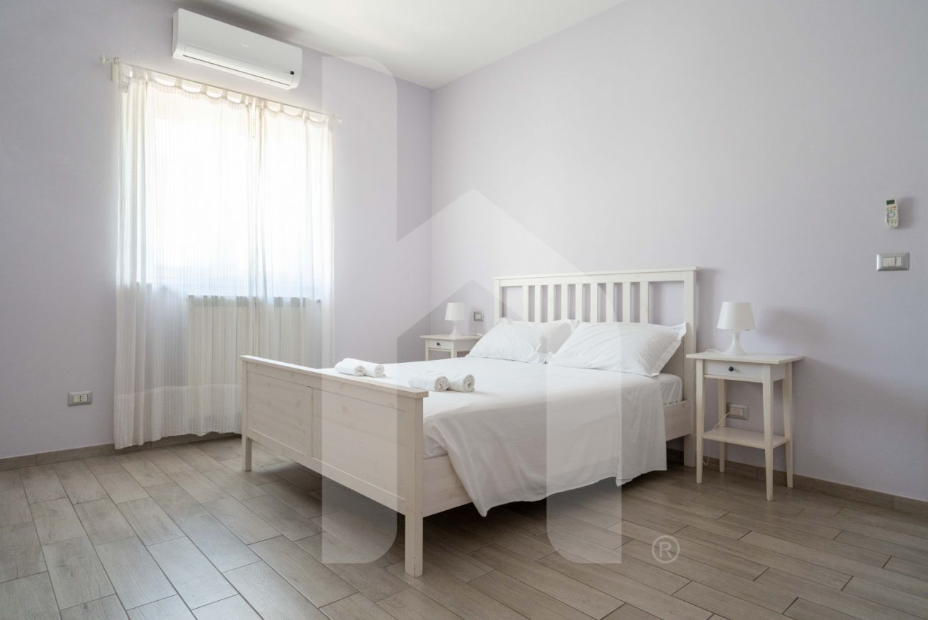 A bedroom with a bed, bedside tables, a window with curtains, and a wall-mounted air conditioner.