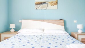 A bedroom with a wooden headboard, a white bedspread with blue floral patterns, and a beach scene painting on the wall. - Mykonos B7 - Apollo - 2-room apartment with terrace and sea view (Pineto)