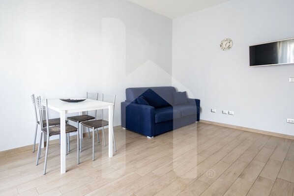Een moderne woonkamer met een witte eettafel, vier stoelen, een blauwe bank en een flatscreen televisie aan de muur.