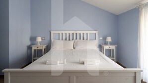 Ein ordentlich bezogenes Bett in einem Zimmer mit hellblauen Wänden, zwei Nachttische mit Lampen und ein Fenster mit Vorhängen.