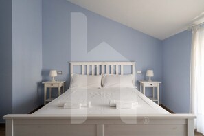 Een net opgemaakt bed in een kamer met lichtblauwe muren, twee nachtkastjes met lampen en een raam met gordijnen.