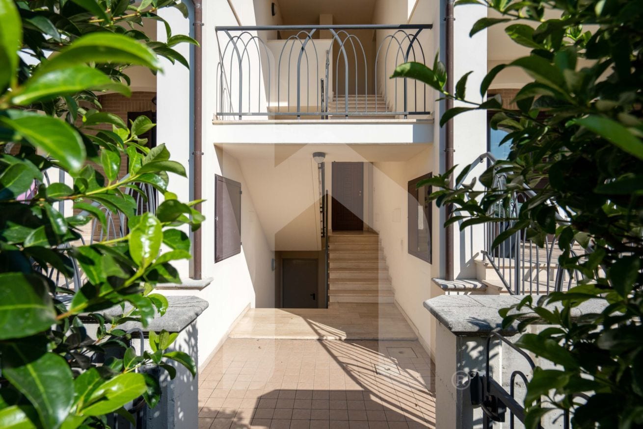 Un edificio residenziale a più piani con una scala centrale, balconi e vegetazione circostante.