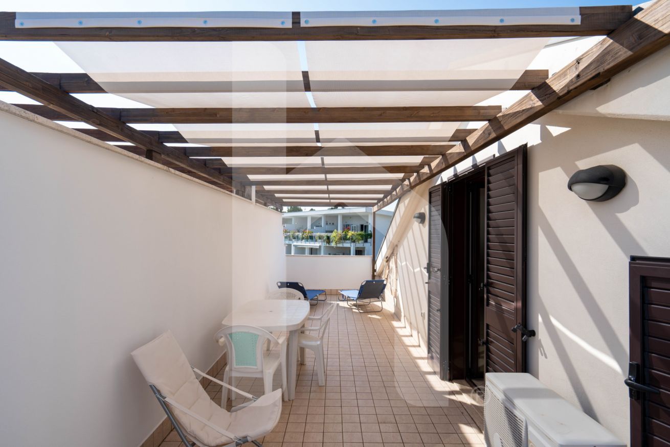 Veranda coperta con tavolo e sedie, vista sulla strada e ingresso di un edificio.