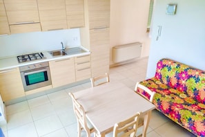 A compact kitchen with a gas stove, oven, and sink, a wooden dining table with chairs, and a colorful sofa.